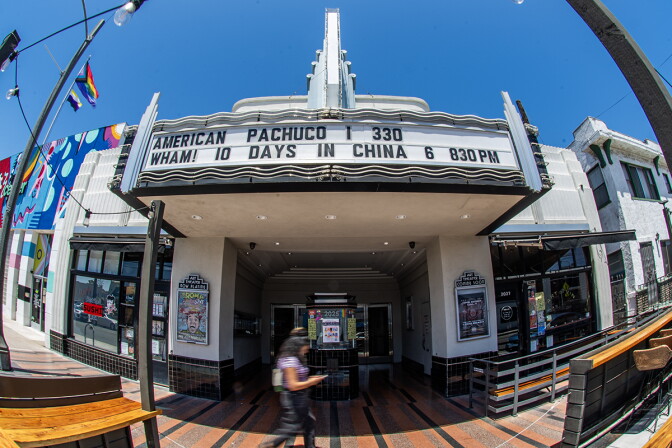 A fish-eye lens view of a person walking past a small theater with signage that reads "American Pachuco | 330. Wham! 10 Days in China 6 830PM."