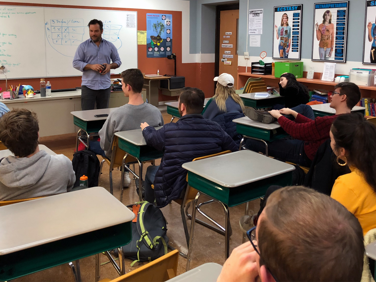 A man in a light blue button-down shirt stands at the front of a high school classroom speaking to students seated at green desks, with a whiteboard, drug-education posters, and "Classroom Rules" signage visible on the walls behind him.
