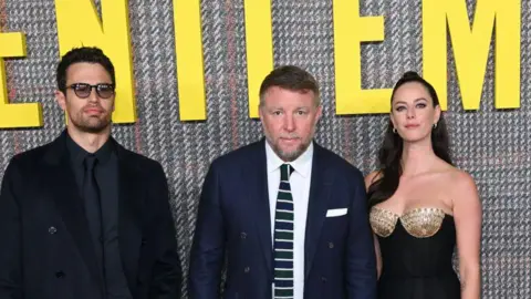 Karwai Tang/WireImage Theo James, Guy Ritchie and Kaya Scodelario pictured at the UK premiere of The Gentlemen in London in 2024. Guy Ritchie is dressed in a dark blue suit, white shirt and a tie featuring white stripes.