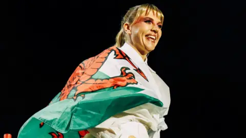 DEPOT LIVE Self Esteem, also known as Rebecca Lucy Taylor, performing on stage at Cardiff Castle. She is wrapped in a Welsh flag, covering her upper body. Her blonde hair is tied up, with her fringe hanging over her forehead. She is smiling and looking away from the camera.