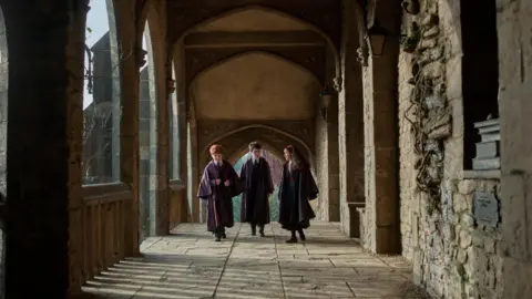 Aidan Monaghan/HBO Max/PA Wire Alastair Stout as Ron Weasley, Dominic McLaughlin as Harry Potter and Arabella Stanton as Hermione Granger walk the halls of Hogwarts in their wizarding robes in HBO Max's new Harry Potter series.
