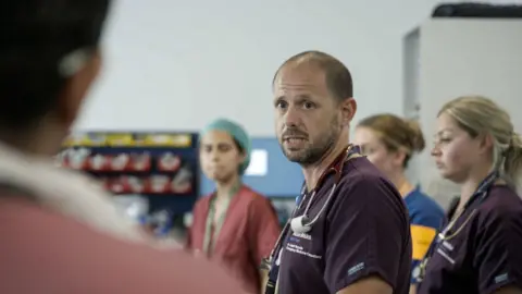BBC/Expectation TV A male nurse captured in a still from the BBC series. He is looking at a colleague with a serious expression on his face, as several staff members stand around at a team briefing.