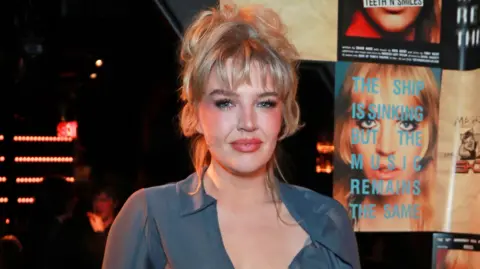Getty Images Self Esteem, also known as Rebecca Lucy Taylor, is standing infront of posters she is pictured in from the play Teeth 'n' Smiles. She is smiling and looking at the camera. Her blonde hair is tied up, with her fringe hanging over her forehead. She is wearing a sheer blue shirt.