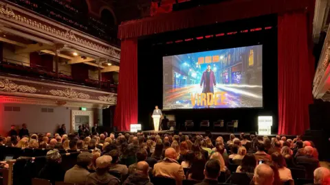 A large screen inside a Victorian theatre with a crowd of people looking on