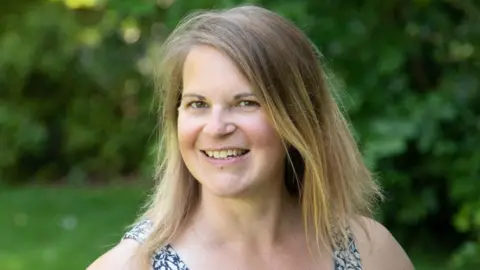 Dr Kathryn Cooper A woman with short blonde hair and brown eyes stands in a garden. She wears a blue and white floral vest top, with blue buttons down the centre. Behind her, grass and leafy bushes can be seen.