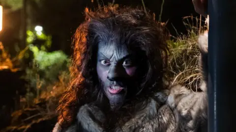 Scott Godding An actress in prosthetic makeup to make her look like a wolf. She wears a wig of long dark hair that has been teased and made to look big and bushy.