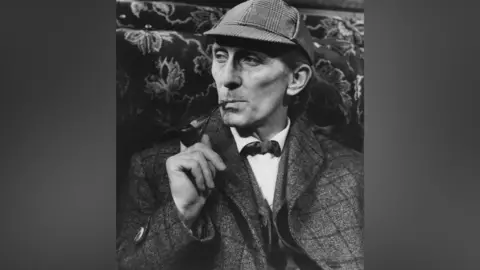 Getty Images A black and white photo of a man in a cap and tweed jacket. He is smoking a pipe.