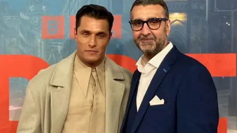 BBC A man in a light-coloured coat and shirt standing next to another man in glasses and a blue suit jacket