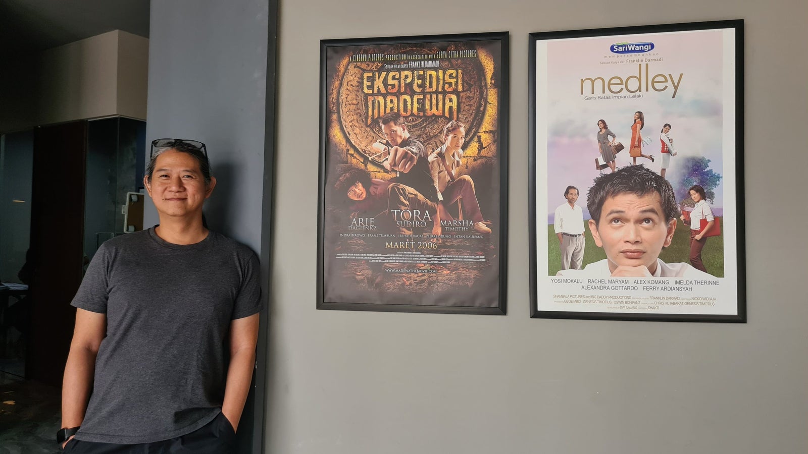 A man with long hair wearing a dark t-shirt and black pants stands next to two movie posters on a wall. The left poster is for the film 'Ekspeadu Madewa,' while the right poster is titled 'Medley,' featuring several people in varied clothing.