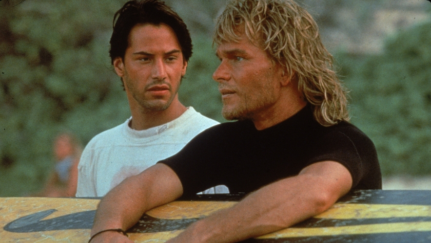 ohnny Utah (Keanu Reeves) and Bodhi (Patrick Swayze) lean on a surfboard together in Point Break
