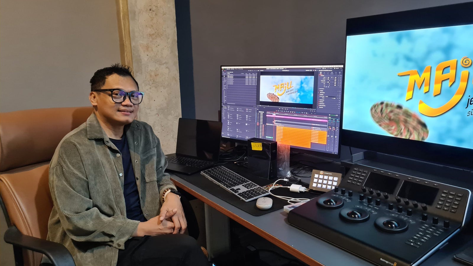 A person sitting in a comfortable office chair in front of a dual-monitor setup, one displaying a colorful graphic with the word 'MAJU' and a turtle design. The person is wearing glasses and a casual outfit, with a desktop featuring editing equipment and a laptop.