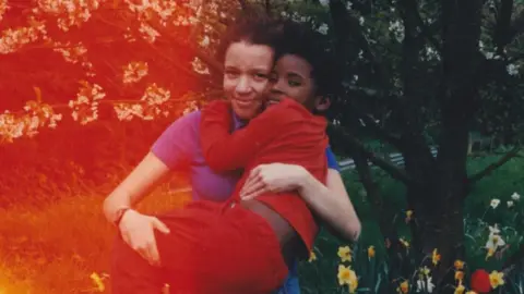 Leah McAleer An older child carries a younger child in a flowering garden, surrounded by blossom-covered trees and spring flowers.  Warm red and orange light leaks across the image, giving the photograph a nostalgic, film-like atmosphere.