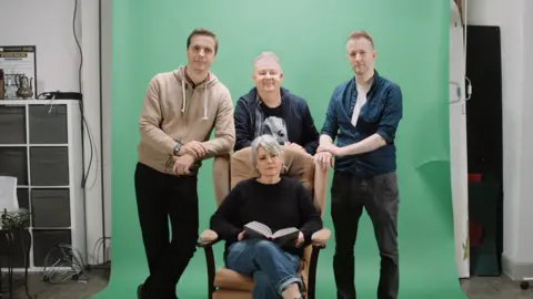 FILM Suffolk Four men stand in front of a woman who is sitting in an armchair. They are smiling at the camera and standing in front of a green screen.