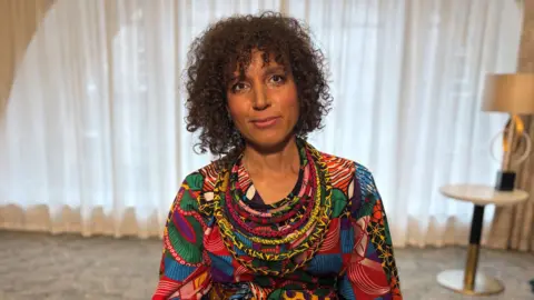 A person wearing a brightly patterned, multicoloured outfit sits indoors against a backdrop of soft curtains and neutral décor.  The vivid clothing stands out against the muted room, creating a striking portrait with warm, natural lighting.