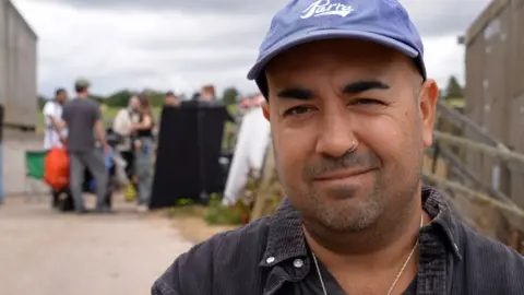Daniel Patrick Vaughan smiles at the camera on the set of MacDonald, which is being filmed on a farm near Hexham. He is wearing a black shirt with a blue hat, silver necklace and silver nose ring. Behind him are multiple people who make up the crew of MacDonald.