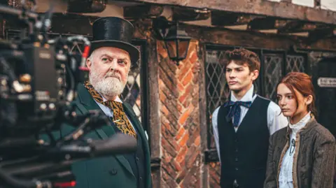 Nick Cooper A behind the scenes image from a film set. A camera on the left hand side of the image pointed toward three actors. One actor wears a top hat and green suit. Another has short red hair and wears a dark waistcoat with a white shirt underneath. The other actress has red hair that has been tied back and she wears a brown jacket with a white blouse underneath.