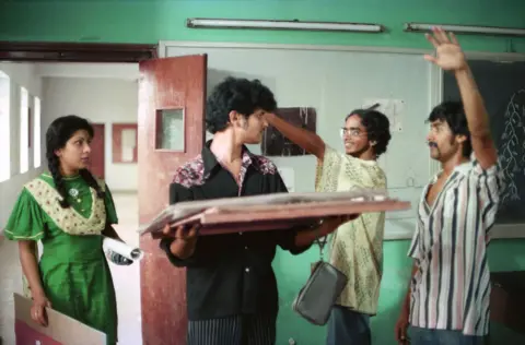 The Film Heritage Foundation Four students - a woman in a green tunic along with three men, dressed in black, yellow and a striped shirt, respectively - stand in a classroom in a scene of the film.