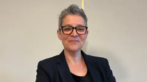 Lou Lockwood a woman with short grey hair smiles against a white background, folding her arms and wearing a dark blazer and T-shirt.