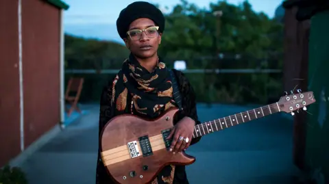Andrew Cawley Bel Odenyo stands outdoors holding a wooden electric guitar, wearing glasses, a dark hat, and a patterned scarf, centered in the frame.
 The scene is shot at dusk with a blurred background of buildings and trees, creating a calm, portrait-style composition.