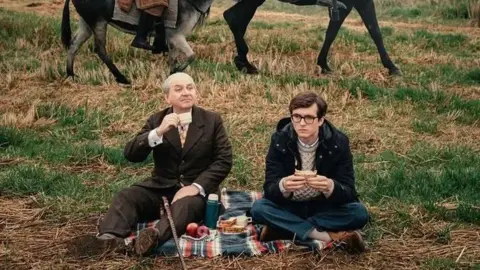 EIFF 2026 Luis Gnecco in brown suit and Fionn Whitehead in dark duffel coat are having a picnic, sitting on a rug in a field. The lower legs of two donkeys can be seen in the background.