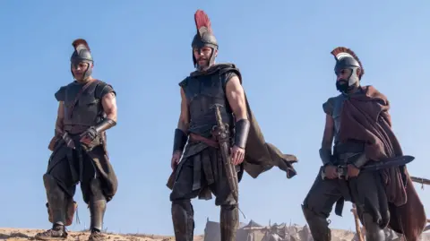 Universal Pictures Three men in costume armour, including helmets with a crest and swords at their hips stand on a mound of sand