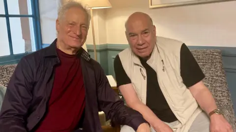 Marc Turtletaub has white hair and is wearing a brown shirt and maroon T-shirt. He is sitting on a chair in a well-appointed room. He is sitting next to Jay Parini, a bald man in a black T-shirt and cream gilet. They are smiling.