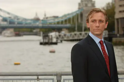 Getty Images On the right of the frame, Daniel Craig stands wearing a suit on a bridge with the London skyline behind him