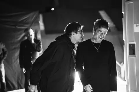 PA Media Liam and Noel Gallagher backstage during their 2025 reunion tour. Liam has on a trademark black cagoule and is saying something to Noel, who is laughing.