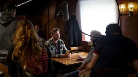 Lewis Brown A behind-the-scenes view of a film or video production inside a wood-panelled pub, as  a man sits at a table holding a drink while crew members and equipment surround the scene.