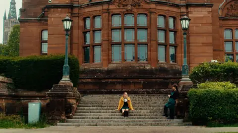 Sonorous Films A wide view of a grand red sandstone building - the Kelvingrove Art Gallery - with large bay windows and ornate architectural details. Broad stone steps lead up to the entrance, framed by decorative lamp posts and neatly trimmed hedges. A woman sits on the steps wearing a bright yellow coat, while another woman stands nearby