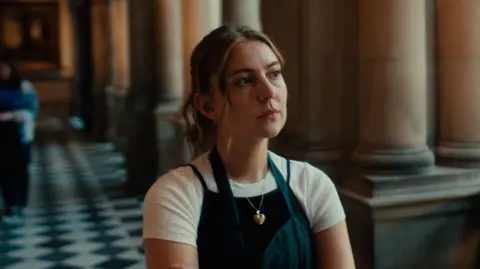 Sonorous Films A woman wearing a dark apron and with light hair over a light-coloured shirt stands inside a grand historic building with tall stone columns and a black-and-white chequered floor. She has a serious expression from her face.