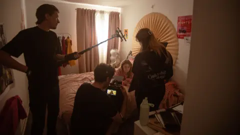 Thomas Buchmann Behind-the-scenes view of a film production taking place in a small bedroom. A woman sits on a bed looking at a mobile phone while crew members surround her