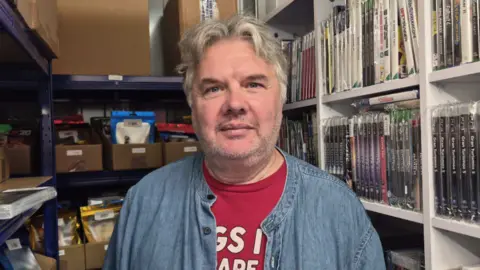 An older man in a red t-shirt and denim unbuttoned shirt looking at the camera, standing in from of boxes and a shelf of video game cases.