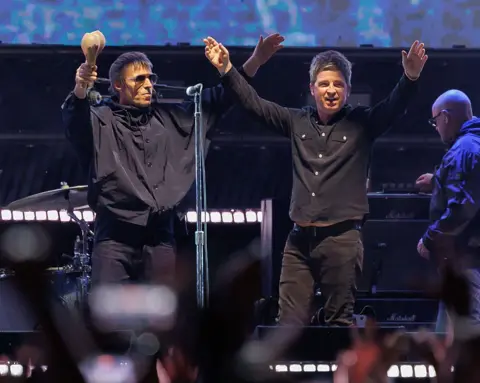 Getty Images Oasis on stage in 2025. Both Liam and Noel are wearing black shirts and raising their arms into the air to greet fans. Noel is holding a pair of maracas in his right hand.