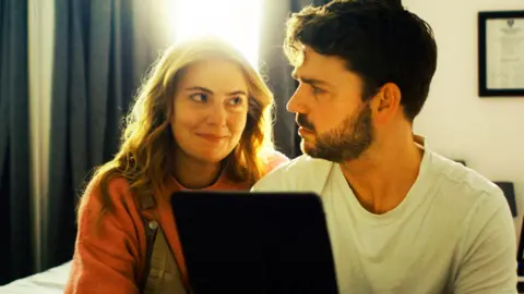 Sonorous Films A man and woman sit closely together in a softly lit room, looking at each other over the top of an open laptop. Bright daylight streams through a window behind them, framed by dark curtains. The woman is wearing a pink jumper and dungarees, while the man is wearing a white T-shirt and has dark hair and a beard.
