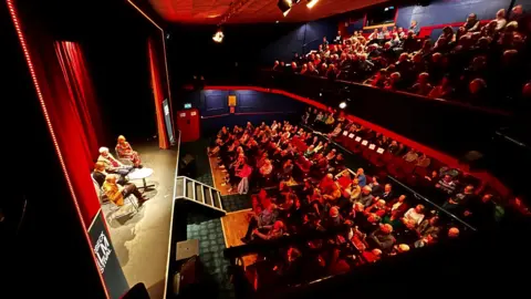 Purbeck Film Festival A view of an event at the Mowlem theatre from a previous Purbeck Film Festival. Red lights are cast on the audience. There are four speakers sat around a small table on the stage.