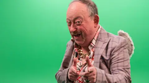 Four Horses Films A man with a moustache wears a suit jacket with an artsy shirt. He wears fairy wings and and a largegold chain necklace. He is pointing at the camera with a slightly threatening smile on his face. The background is a green screen.