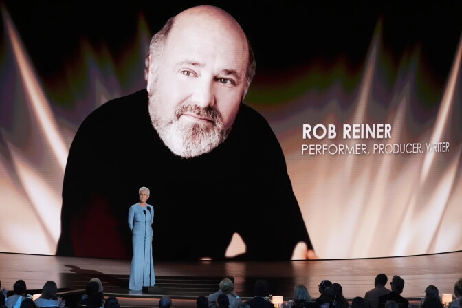 Jamie Lee Curtis, a woman wearing a blue dress, stands on a stage in front of a screen showing a photo of Rob Reiner as she faces an audience sitting down.