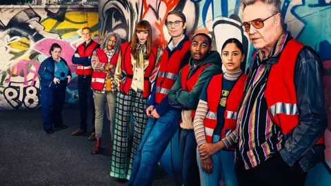 BBC The cast of The Outlaws lined up against a graffitied wall, wearing red hi-vis vests