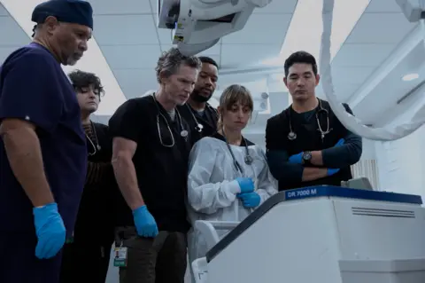 Warner Brothers A group of medical staff gather round a hospital machine