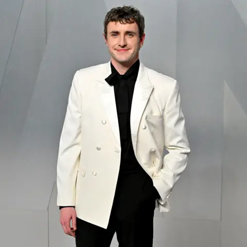 Getty Images Paul Mescal smiles while posing for a photo wearing a white tuxedo with black shirt and trousers against a grey background, in LA in March.