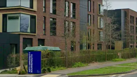 Google The exterior of a red-brick and black building with many windows on the left hand side of a road. A tree-lined fence separates the road and the building. A dark blue sign between the pavement and the building is reads "ARTS UNIVERSITY BOURNEMOUTH".