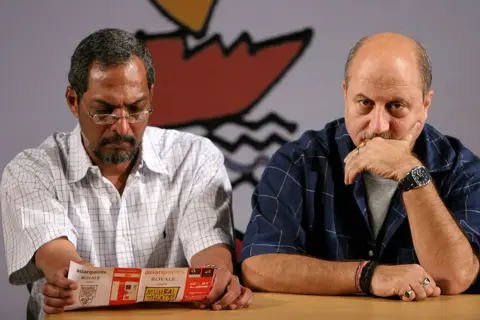 Getty Images Shot of Nana Patekar and Anupam Kher sitting on a table on stage as part of a performance in a play in 2006. Patekar is wearing a white checked shirt with glasses looking at a pamphlet while Kher is wearing a blue checked shirt looking off-camera resting his head in his left hand.